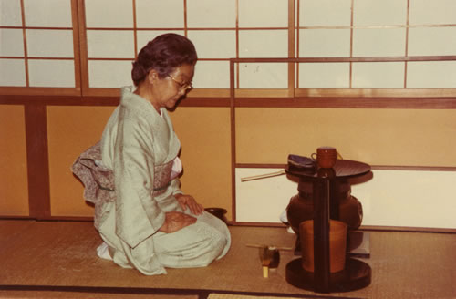 Woman in kimono kneeling in front of tea kettle placed over a heater, tea cup, and whisk.